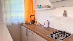Et køkken med gaskomfur, vask og et vindue med let gardin og orange stofgardin.