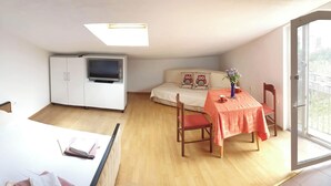 Ett litet, ljust rum med en säng, en soffa, ett matbord med stolar, en tv och en balkong.