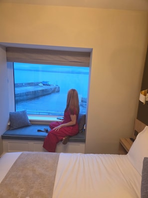 A room with a bed, a window seat with a view of the sea, and a bedside table with a lamp.