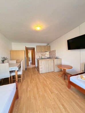 Ett kompakt hotellrum med trägolv, en matplats med bord och stolar, ett litet kök, en säng och en tv som är monterad på väggen.