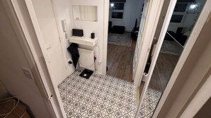 Um corredor estreito com piso estampado, uma porta branca, um pequeno estante branco com um telefone e um alto-falante, e um espelho na parede.