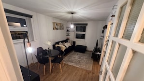 Sala de estar com um sofá preto, uma mesa redonda e uma mesa de centro. Há um candelabro pendurado no teto.