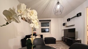 Uma sala de estar moderna com uma mesa de jantar, cadeiras pretas, uma planta em vaso, uma TV e uma luminária geométrica.