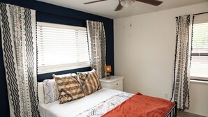 Un dormitorio con cama, cortinas, una mesita de noche, lámpara y ventilador de techo.