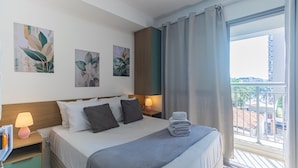 Camera d'albergo con un letto, cuscini, comodino con lampada, opere d'arte al muro e vista sulla città attraverso una finestra.