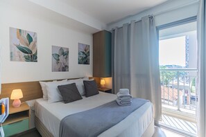Ein Hotelzimmer mit Bett, Kissen, einem Nachttisch mit Lampe, einem Kunstwerk an der Wand und Blick auf die Stadt durch ein Fenster.