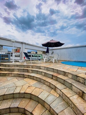A rooftop pool area with steps leading to the pool, white lounge chairs, and a large umbrella. - Duplex w/ Pool & BBQ 500m to Enseada Beach (Guarujá)