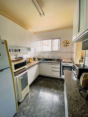 A kitchen with white cabinets, a microwave, an oven, a fan, and a window with blinds. - Duplex w/ Pool & BBQ 500m to Enseada Beach (Guarujá)