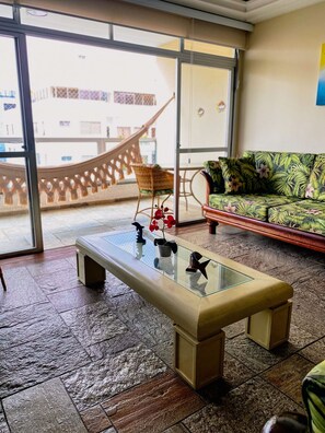 A room with a glass-top coffee table, a wicker chair, a sofa with a floral pattern, and a view of a building through a glass door. - Duplex w/ Pool & BBQ 500m to Enseada Beach (Guarujá)