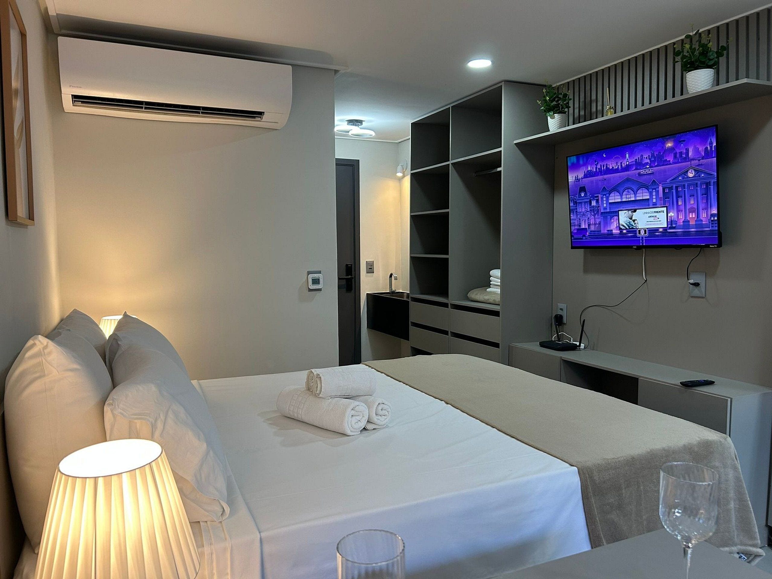 A hotel room with a bed, a TV mounted on the wall, a bedside table with a lamp, and a glass of water.