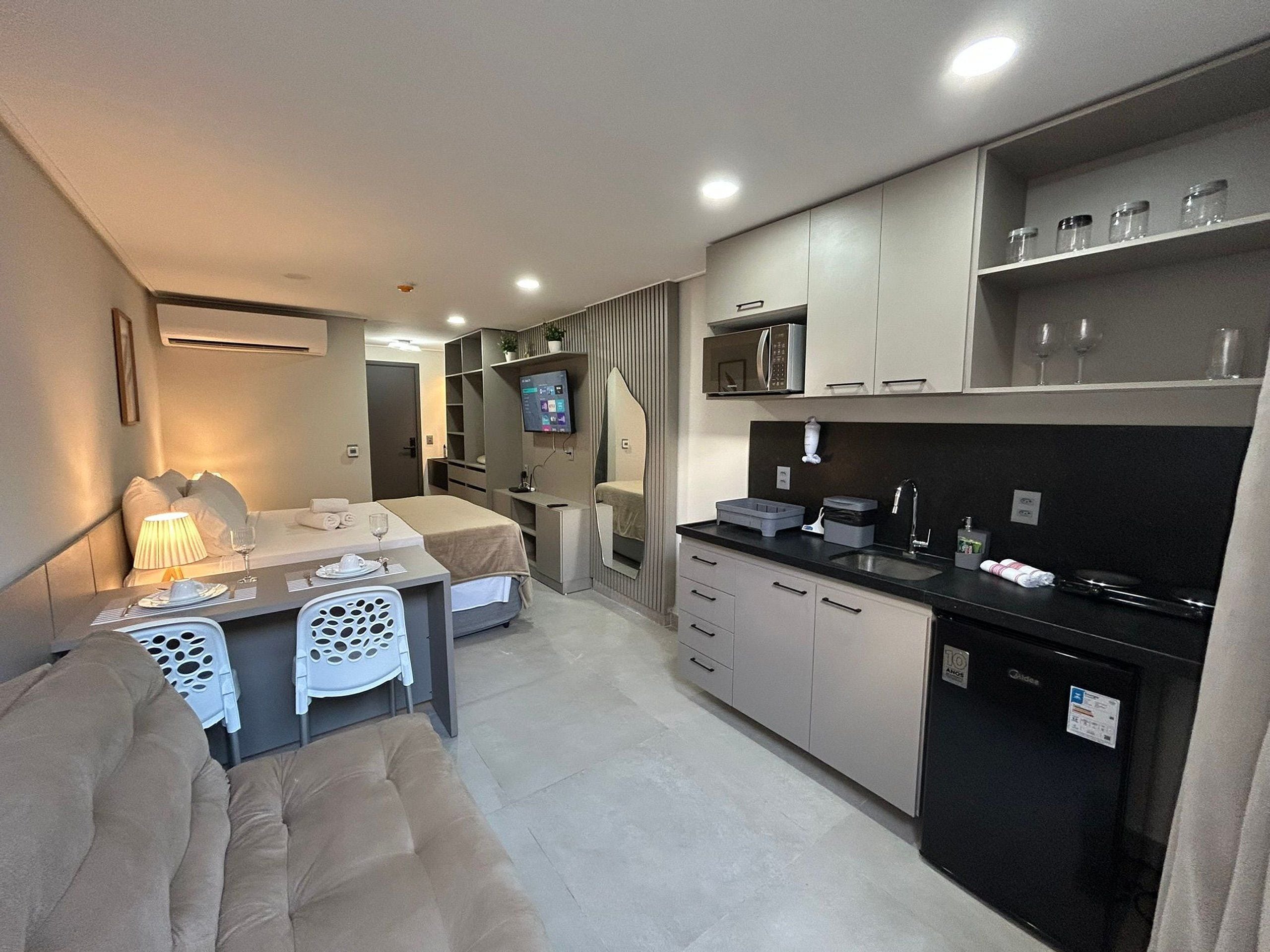 A compact hotel room with a kitchenette, a dining area with a table and chairs, a bed with a headboard, and a TV mounted on the wall.