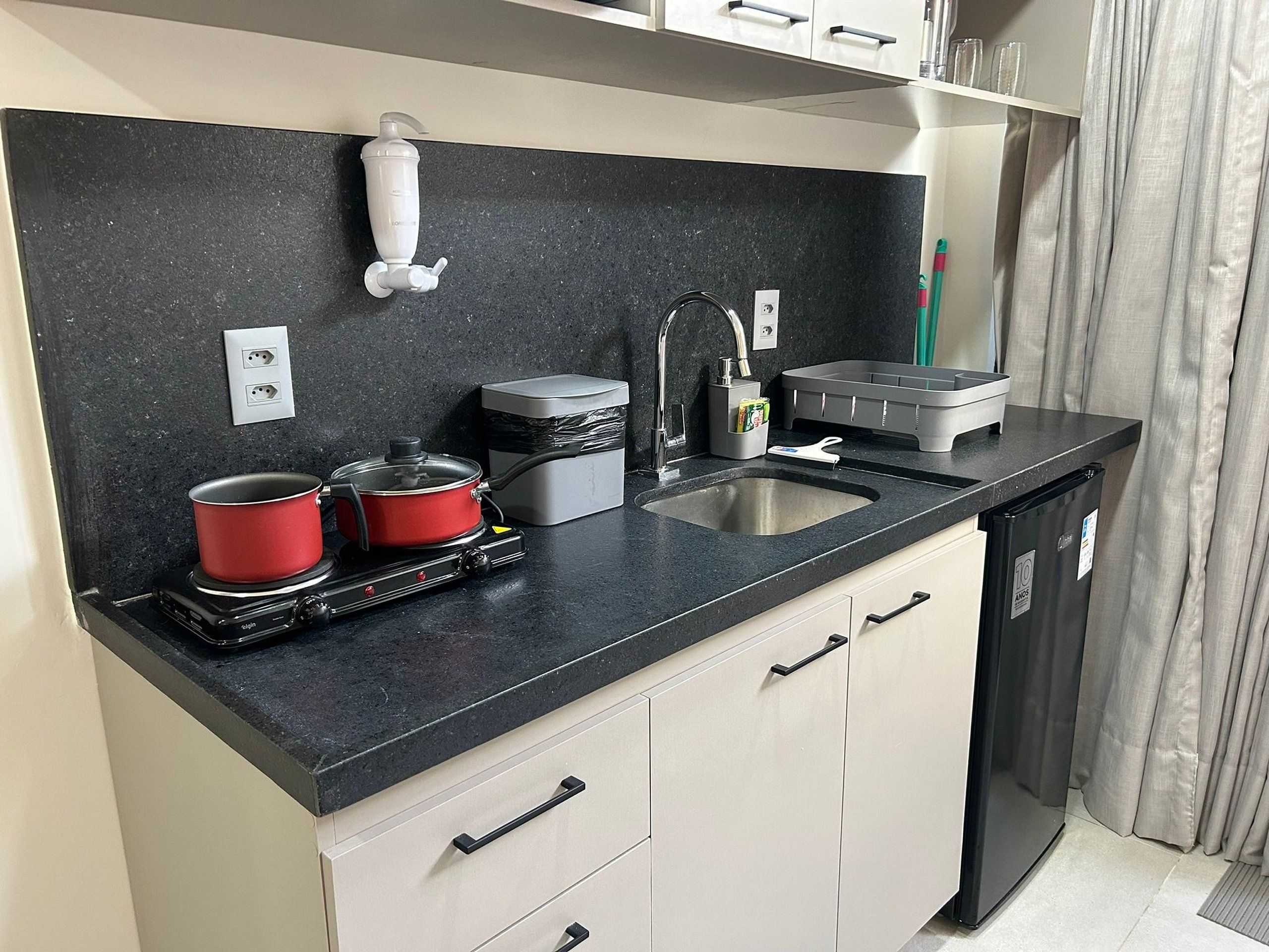 A compact kitchen with a black countertop, two red pots, a small stove, a sink, and a compact refrigerator.