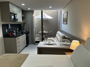 Ein kompaktes Hotelzimmer mit Küchenzeile, einer Essecke mit Tisch für zwei Personen, einer Couch und einem großen Fenster mit Sonnenschirm.
