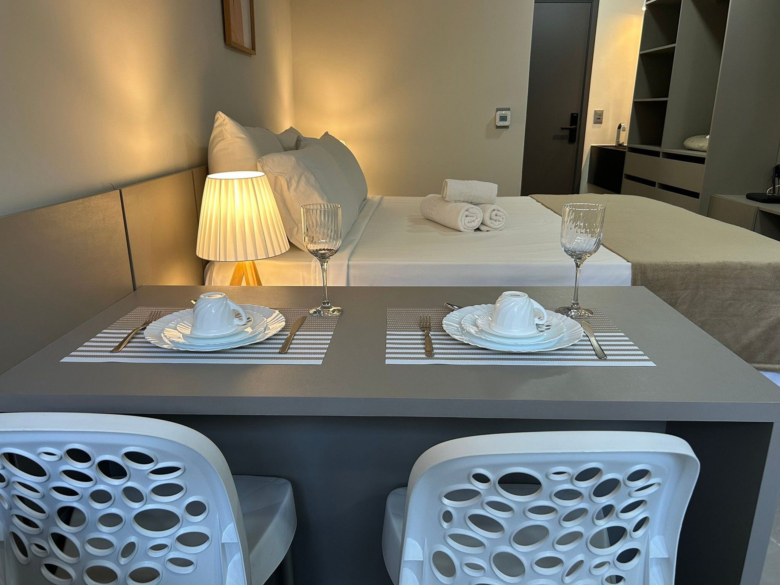 Ein Hotelzimmer mit einem ordentlich bezogenen Bett, einem Esstisch gedeckt mit Tellern, Gläsern und Besteck sowie zwei Stühlen mit Löchern.