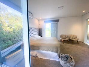 Ein Schlafzimmer mit einem großen Bett, zwei Stühlen, einem Platz für ein Haustier und Blick auf Bäume durch eine Glastür.