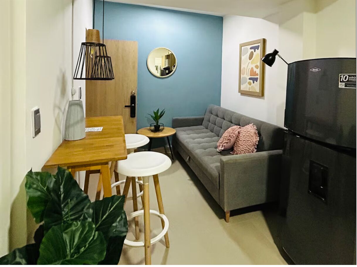 Un espacio habitable pequeño con sofá, una mesa de comedor con sillas, una mesita auxiliar, refrigerador, espejo y una planta en maceta.