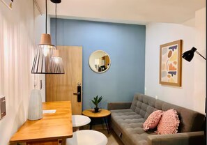 Ein Wohnzimmer mit Holztisch, grauem Sofa, rundem Spiegel und kleinem Tisch mit Pflanze.