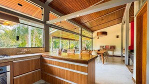 A modern kitchen with wooden cabinetry, a large island, and a dining area with a view of greenery. - Luxury Single-Story House in Granja Viana (Jandira)