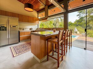 A modern kitchen with wooden cabinetry, a central island with bar stools, a refrigerator, and a view of a pool and greenery outside.
