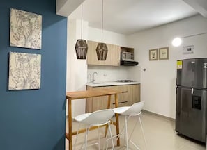 Eine kleine Küche mit einem Holztisch und weißen Stühlen, einem Edelstahlkühlschrank sowie zwei hängenden Pendelleuchten.