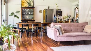 A spacious living area with a wooden floor, a pink sofa, a dining table with chairs, and a kitchen area with a refrigerator and shelves. - Charming & Spacious Home w/ Garden | 2BR (Carapicuíba)