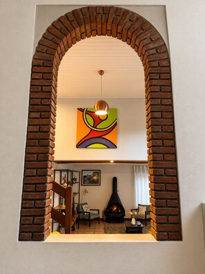 A living room with a brick fireplace, a colorful abstract painting, and a wooden staircase. - Spacious House with Large Pool and Barbecue Near São Paulo (Carapicuíba)