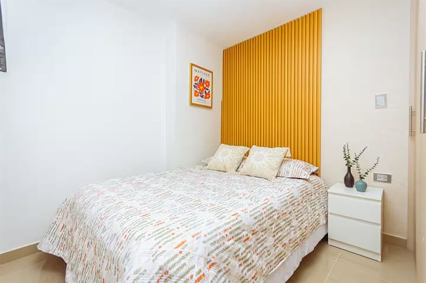 Une chambre à coucher avec un lit, une table de chevet, une étagère fixée au mur et un tableau encadré.