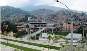 Ein modernes Gebäude mit einer zentralen Treppe, umgeben von einer begrünten Anlage mit Bäumen und freiem Blick auf eine hügelige Stadtlandschaft im Hintergrund.