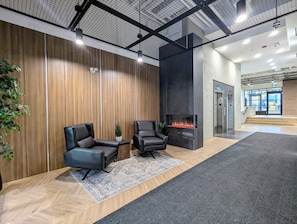 Um hall com duas poltronas de couro, uma mesinha, um vaso de planta, uma lareira e uma escadaria que leva a um elevador.