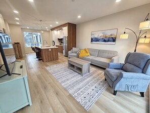 Una sala moderna con sofá, sillón, mesa de centro y una cocina al fondo.