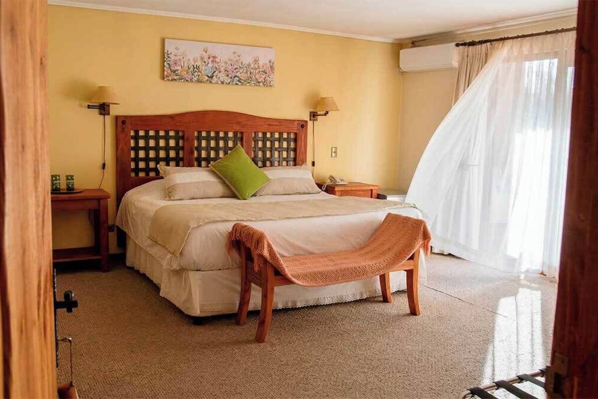 Un dormitorio con cabecera de madera, una cama con almohada verde, una mesita de noche, una lámpara y una ventana con cortinas.