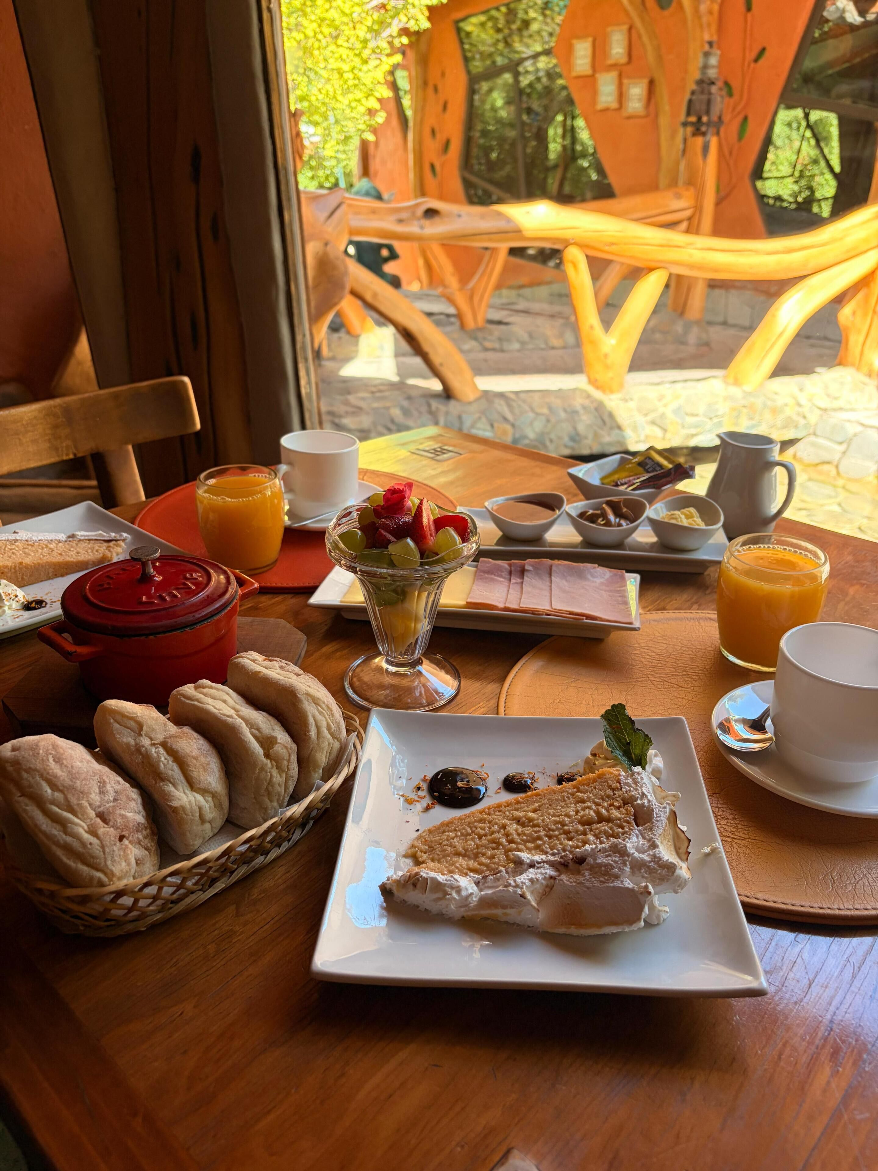 Desayuno con pan, fruta y una rebanada de pastel sobre una mesa.