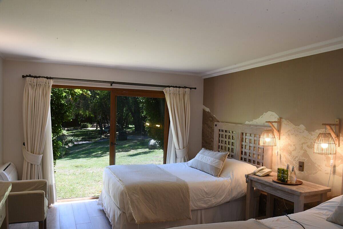 Un dormitorio con un ventanal grande, una cama, una mesita de noche con lámpara y vistas a un jardín.