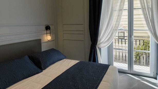 Un dormitorio con cama, luz empotrada en la pared, ventana con cortinas y vista al balcón exterior.