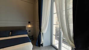 Una habitación de hotel con cama, mesita de noche, lámpara de pared y ventana con cortinas.