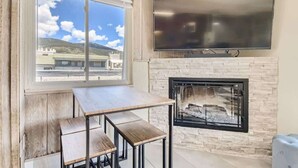 A room with a stone fireplace, a flat-screen TV mounted above it, and a large window offering a view of buildings and mountains. - Amazing Lake and Mountain Views! (Dillon)
