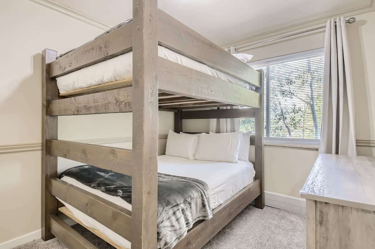 Un letto a castello con biancheria da letto bianca e una coperta grigia, una finestra con persiane e un comodino in legno.