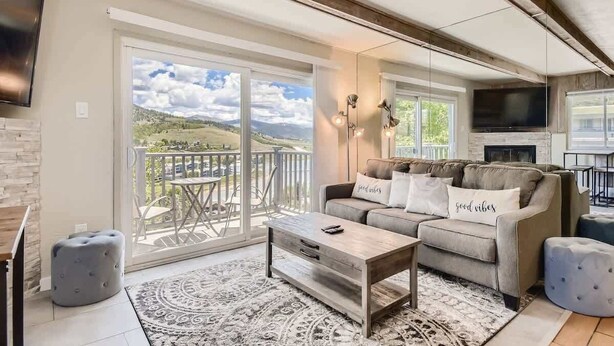 A spacious living room with a large sliding glass door leading to a balcony, a sofa with decorative pillows, a coffee table, and a flat-screen TV mounted on the wall. - Amazing Lake and Mountain Views! (Dillon)