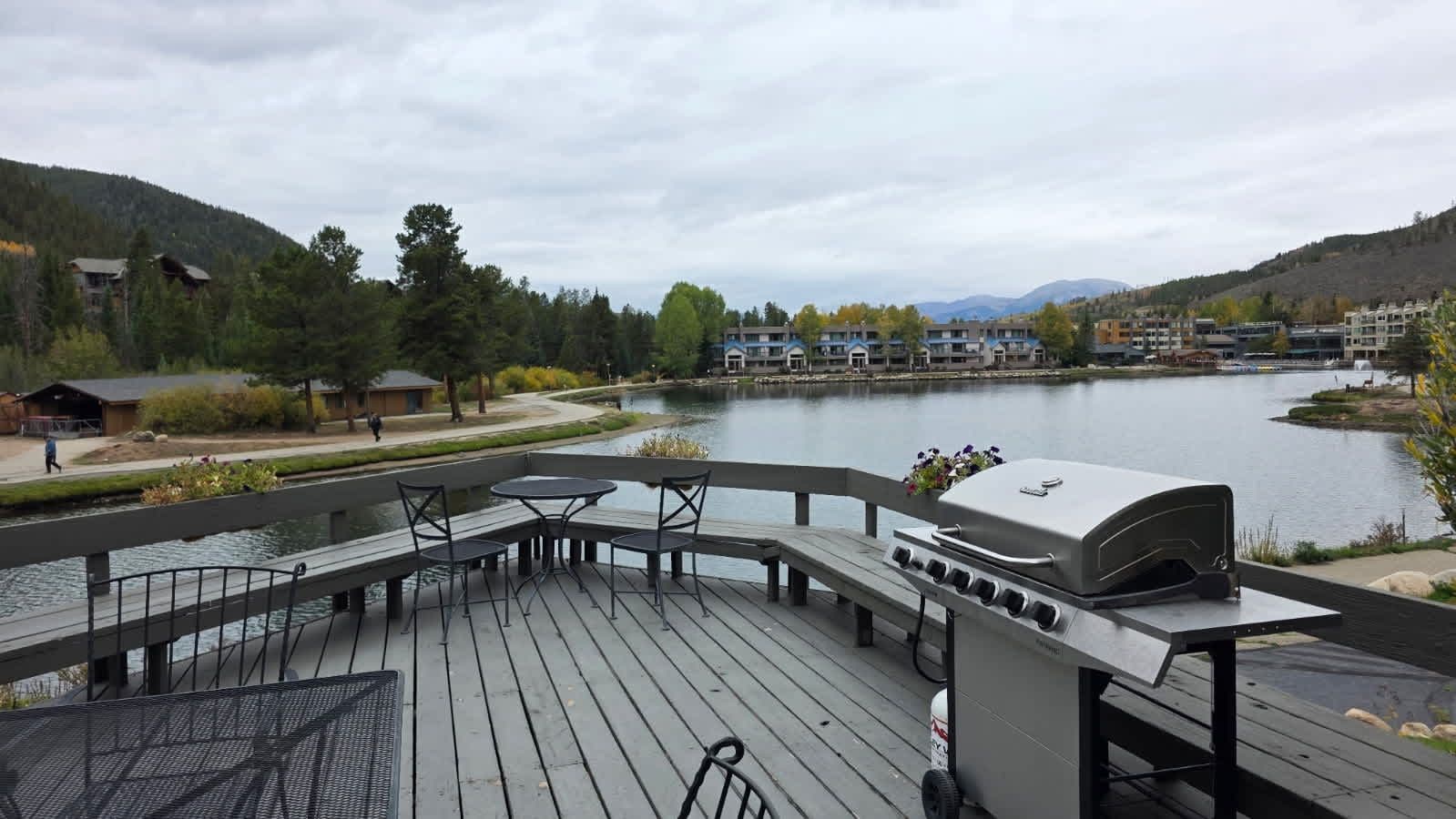 A deck with a grill and seating overlooking a lake, surrounded by buildings and trees.