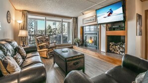A cozy living room with a leather sofa, a wooden coffee table, and a fireplace. A television is mounted on the wall, and there is a large window providing a view of the outdoors. - Close to Slopes, Pool, & Nature: The Pines Condo in Beautiful Keystone (Keystone)
