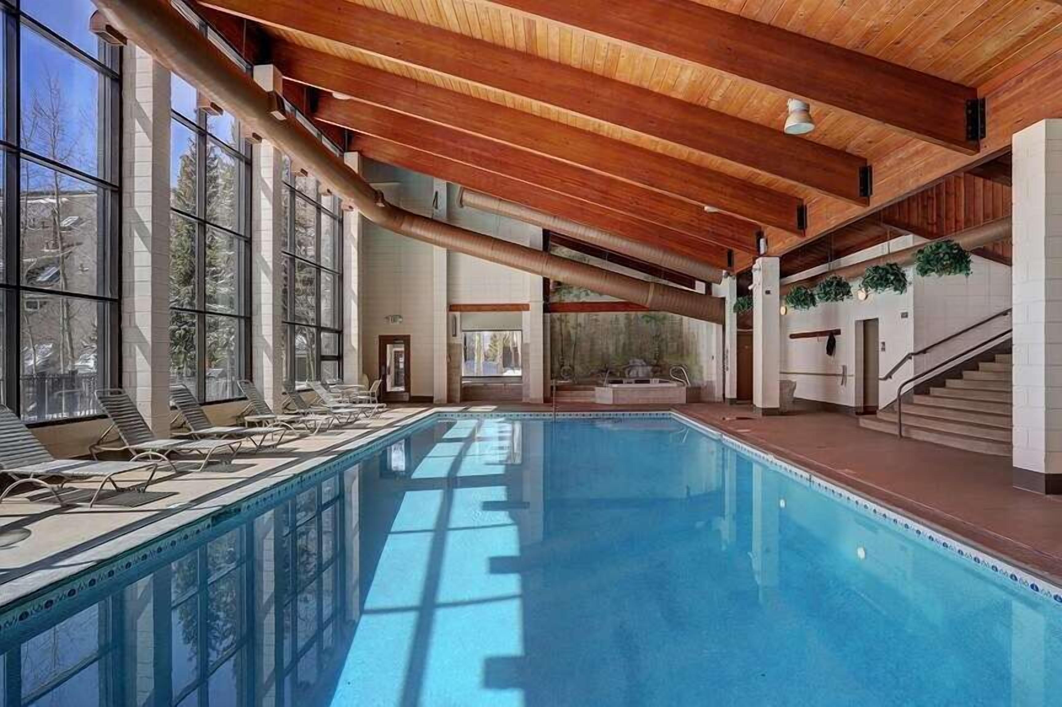 Una piscina interior con ventanas iluminadas por el sol, sillones de descanso y techo de madera.