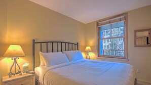 A neatly made bed with white linens, two bedside lamps, a nightstand with a tissue box, and a window with a view of trees. - Heart of River Run Village! (Keystone)