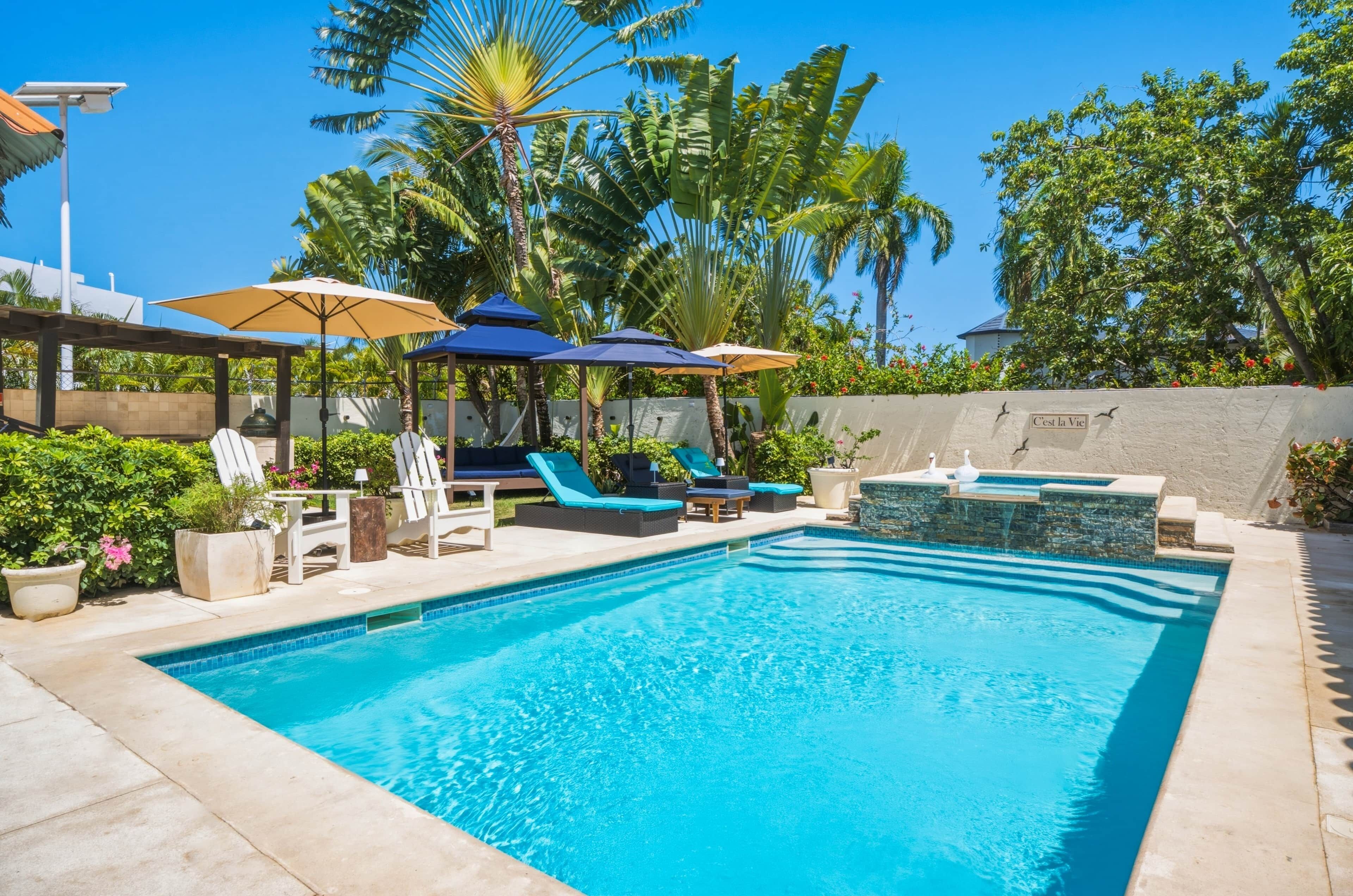 A swimming pool with lounge chairs and umbrellas, surrounded by tropical plants and a stone wall.