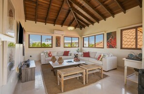 Ein geräumiges Wohnzimmer mit hoher Decke, großen Fenstern, einer weißen Sofagarnitur, einem Couchtisch und einem an der Wand montierten Fernseher.