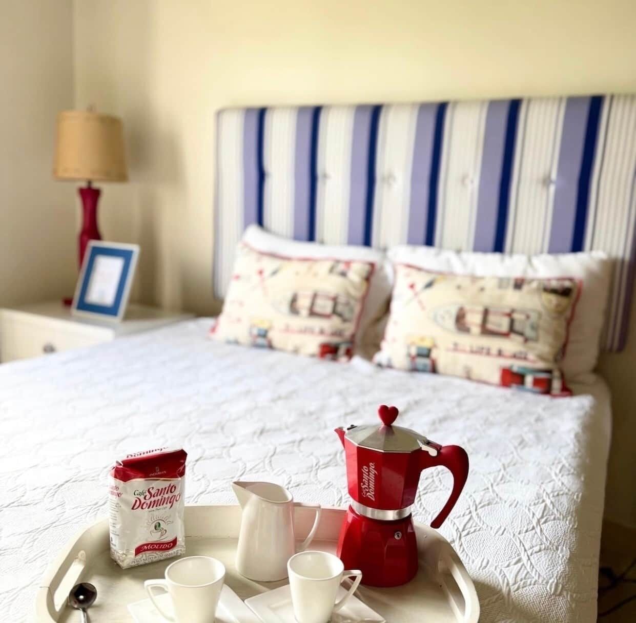 A neatly made bed with a striped headboard, a bedside lamp, and a tray with a coffee pot, cups, and a packet of coffee.