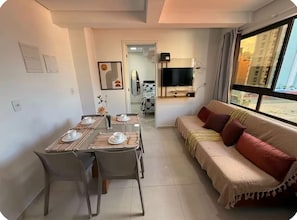 Ein modernes Wohnzimmer mit einer Couch, einem Esstisch für zwei Personen, einem an der Wand montierten Fernseher und einem großen Fenster.