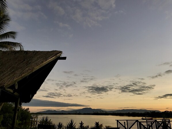 A thatched-roof structure overlooking a calm lake with distant mountains and a cloudy sky.