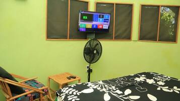 A room with a bed, a chair, a small table, a TV mounted on the wall, and a fan.