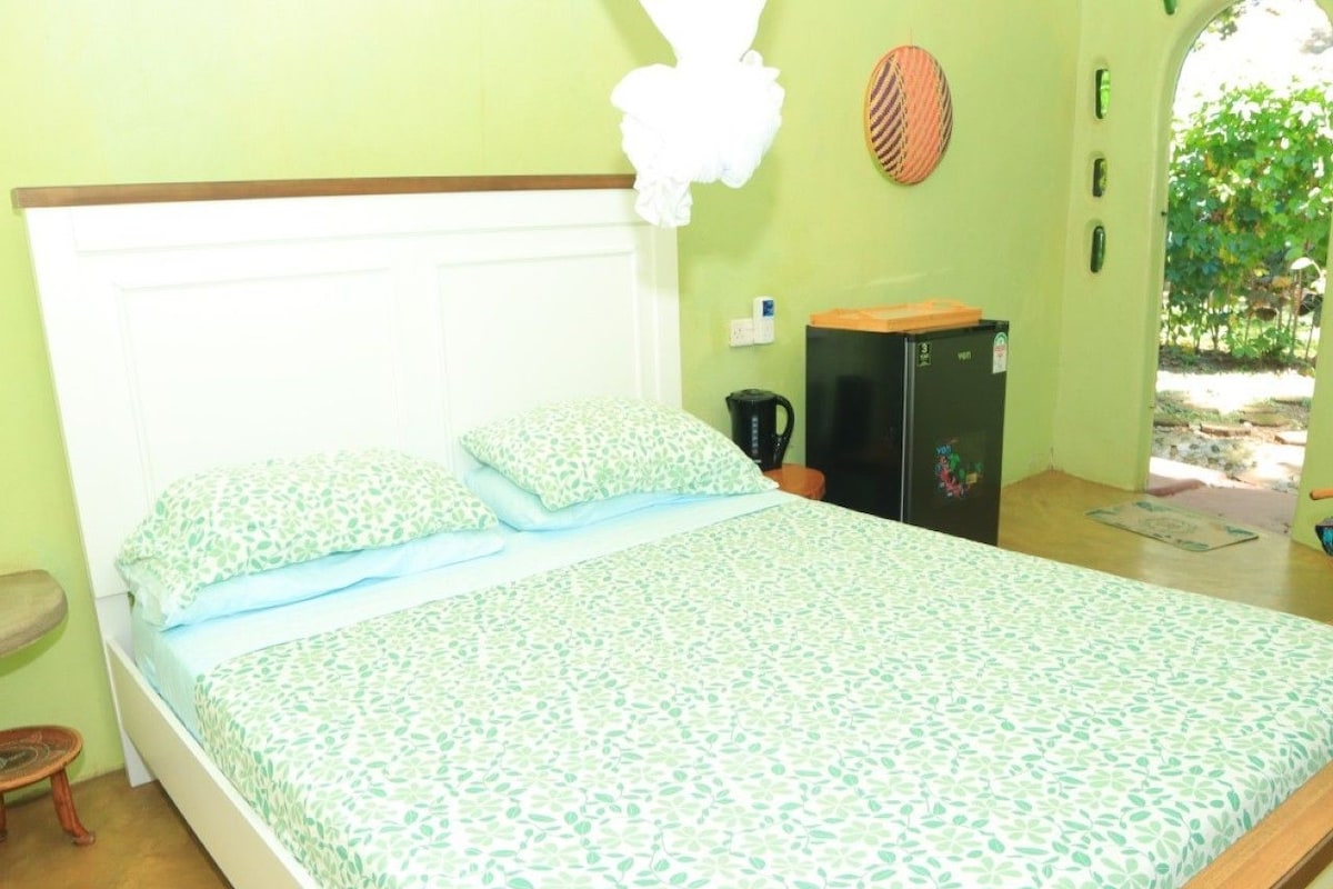 Una cama individual con una colcha estampada, una mesita de noche, una silla, una pequeña mesa, un hervidor y un refrigerador.