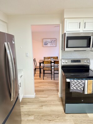Een keuken met een koelkast, magnetron, oven en een houten eettafel met stoelen.